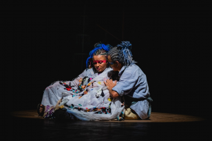 Apresentação de teatro onde duas crianças estão sentadas e uma segura a outra, ambas usam cabelos presos e uma roupa longa azul.